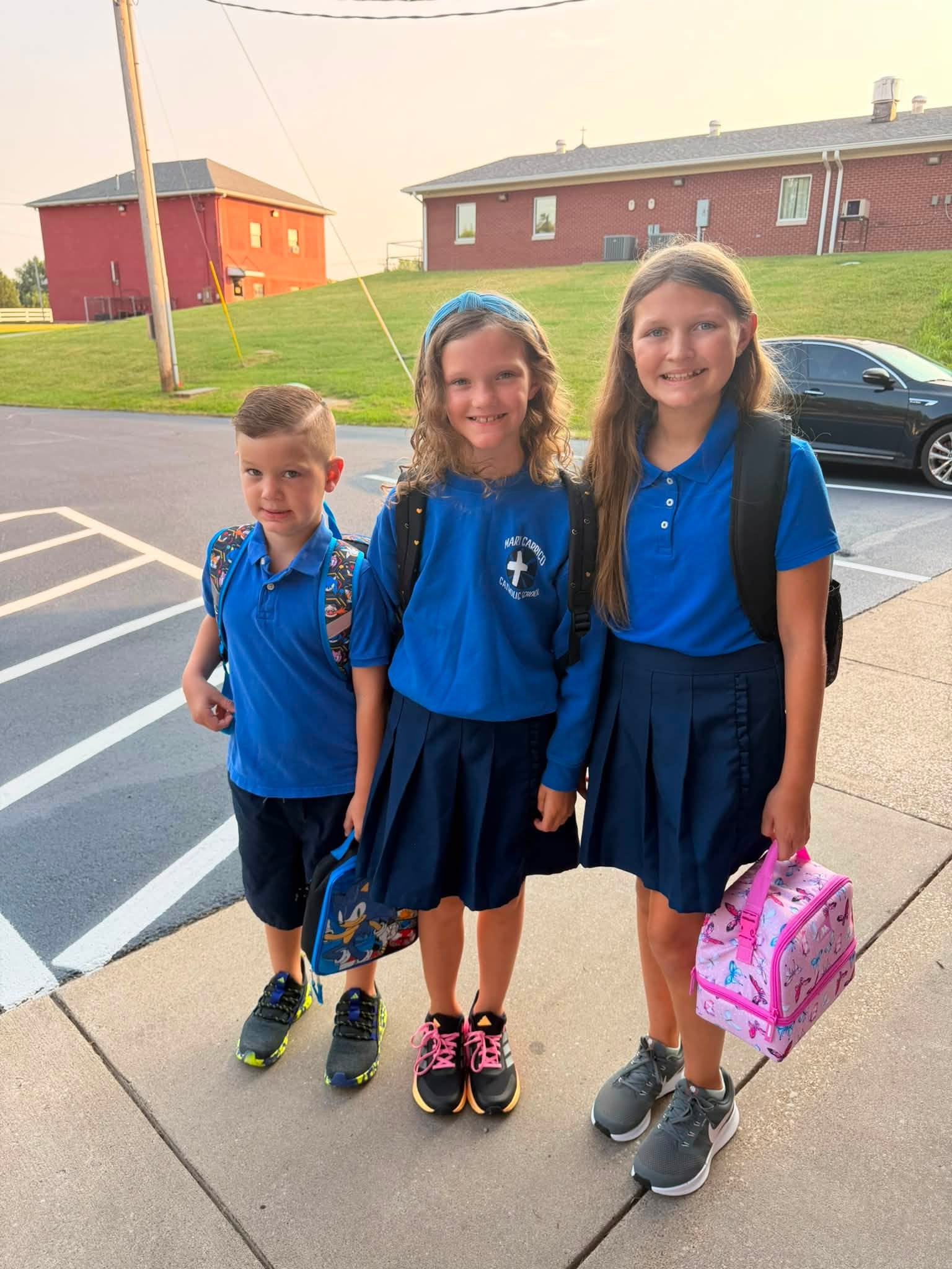 Three students in blue uniforms stand outside with backpacks and lunch bags.