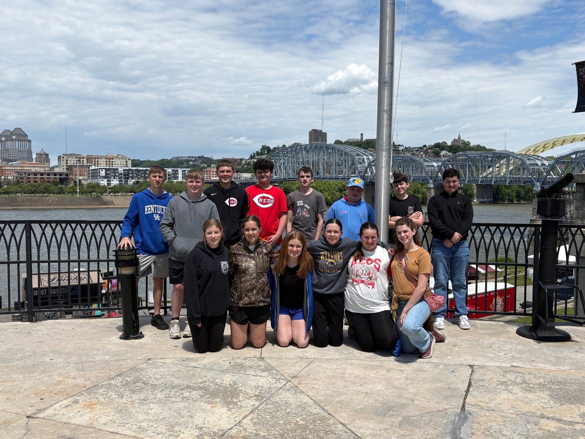 Students gather for a group photo beside a river, with a bridge and city skyline behind them.
